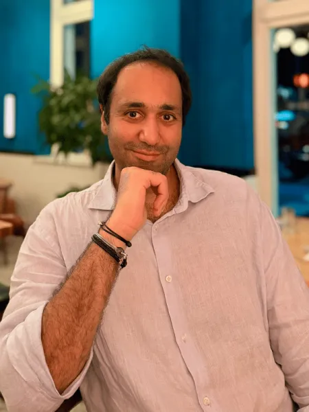 A smiling man in a lavender button-down shirt rests his chin on his hand while seated in a restaurant.